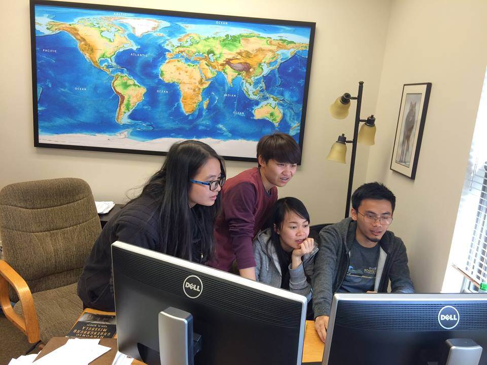 A group of threee fellows looking at a computer screen with a world map behind them